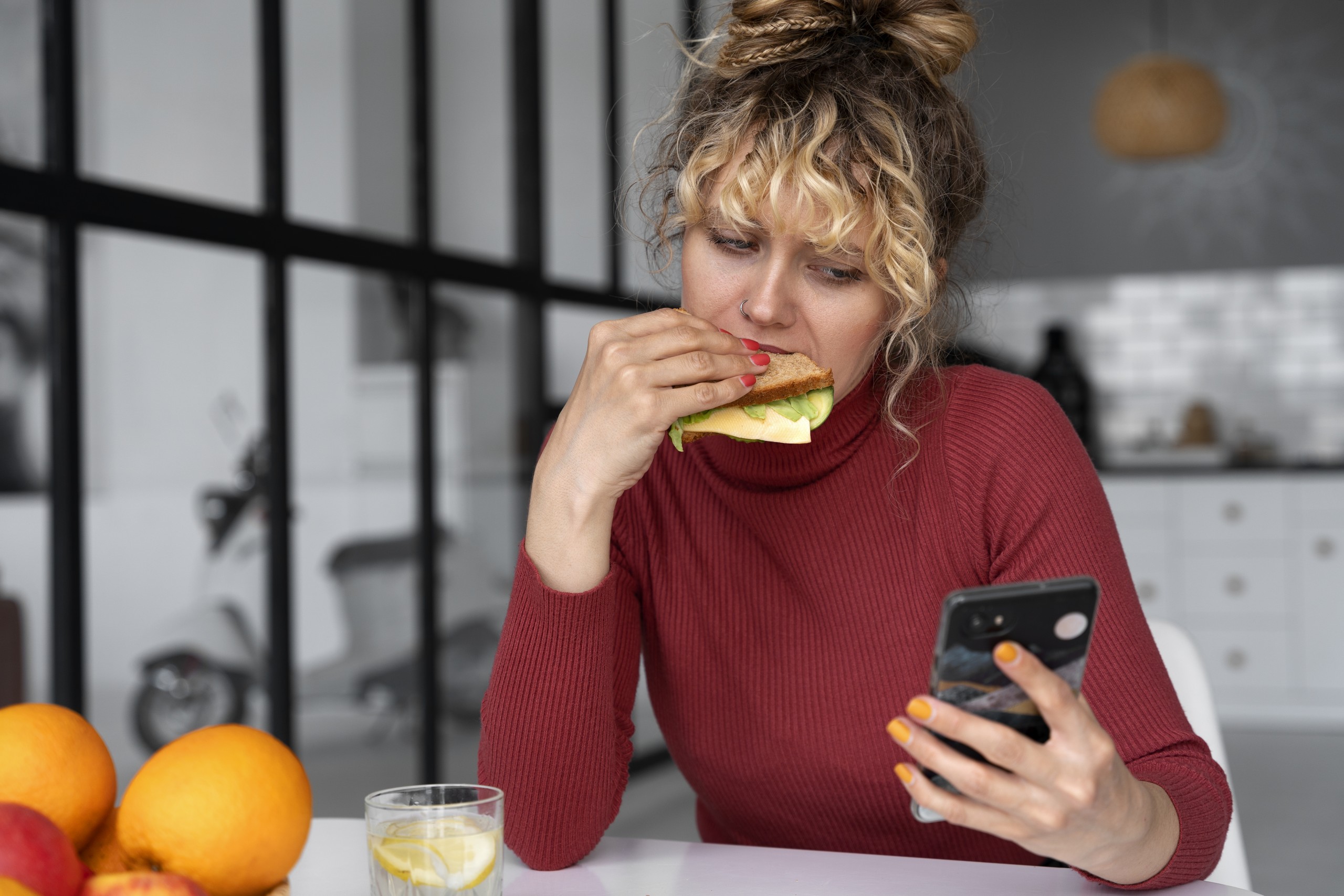 mujer rubia con pelo recogido polera roja mirando su celular mientras come un sándwich.